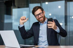 geslaagd volwassen zakenman in kantoor luisteren naar muziek- en dansen, baas in bril met telefoon vieren prestatie werken binnen gebouw gebruik makend van laptop Bij werk. foto