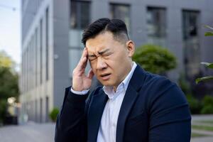 bezorgd Aziatisch zakenman in een blauw pak gevoel een hoofdpijn, staand buiten een kantoor gebouw met een gepijnigd uitdrukking. foto
