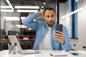 verrast mannetje professioneel in een gewoontjes overhemd looks Bij zijn cel telefoon met een verbaasd uitdrukking. een modern, goed verlicht kantoor ruimte met een laptop en notitieboekje zichtbaar in de achtergrond. foto