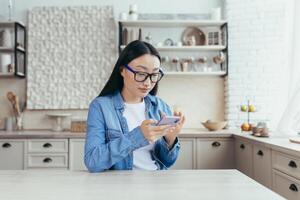 jong mooi Aziatisch vrouw zittend in keuken Bij huis en gebruik makend van mobiel telefoon. scrollt door de nieuws voer, zit Aan sociaal netwerken, cheques mail. foto