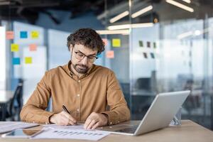 volwassen Mens met bril gefocust Aan werk, herzien documenten en gebruik makend van een laptop in een modern kantoor instelling, omringd door kleverig notities. foto