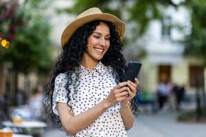 een mooi jong vrouw wandelingen door de avond stad in een hoed, een glimlachen Latijns Amerikaans vrouw houdt een smartphone in haar handen. een toerist met gekruld haar- types een bericht en bladert online Pagina's Aan de telefoon. foto