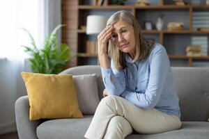 attent ouderen dame zittend met gekruiste benen en resting hoofd Aan hand- met gefrustreerd uitdrukking.moe vrouw lijden van hoofdpijn terwijl wrijven voorhoofd en op zoek vooruit met teleurstelling. foto