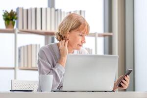 portret van vrouw gebruik makend van smartphone en laptop, volwassen vrouw in leven kamer met mobiel telefoon browsen internet Aan modern computer apparaatje foto