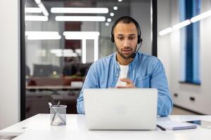 Latijns Amerikaans Mens in een koptelefoon zit in de kantoor in voorkant van een laptop en communiceert Aan een telefoongesprek. foto