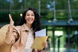 opgewonden zakenvrouw Holding documenten, tonen succes gebaar buitenshuis in een stedelijk instelling. uitdrukkingen van professioneel triomf en geluk. foto