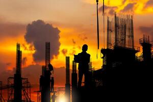 silhouet van ingenieur en arbeider team controle project Bij gebouw plaats achtergrond, een bouw plaats met zonsondergang foto