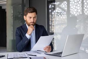 een gefocust mannetje accountant beoordelingen financieel papieren met een echt uitdrukking, diep in gedachte Bij zijn werkplaats met een laptop en rekenmachine Bij hand. foto