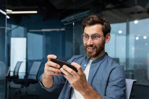 volwassen zakenman met baard is spelen spel Aan smartphone, Mens is hebben pret gedurende breken Bij werkplek, zakenman binnen modern kantoor. foto