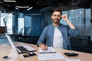 portret van geslaagd financier Bij werk Bij werkplaats binnen kantoor, volwassen Mens glimlachen en op zoek Bij camera, zakenman in overhemd werken met documenten grafieken en grafieken, papierwerk gebruik makend van laptop. foto