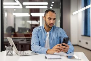 Latijns Amerikaans jong Mens zakenman, freelancer werken in de kantoor, zittend Bij de tafel, ernstig en geconcentreerd gebruik makend van de mobiel telefoon. foto