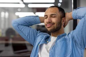 een jong Indisch Mens in denim kleding glimlachen met ogen gesloten, ontspannende in een modern kantoor instelling, uitstralend vrede en tevredenheid. foto