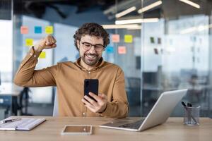 blij Mens Bij werkplaats met een laptop, verhogen vuist terwijl op zoek Bij de smartphone, uitdrukken succes en geluk. foto