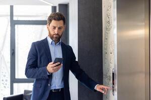 gefocust zakenman in een pak gebruik makend van smartphone terwijl aan het wachten voor een lift in een modern kantoor gebouw interieur. foto