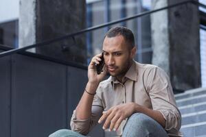 depressief Mens zittend Aan trap van kantoor gebouw, gefrustreerd pratend Aan telefoon, van streek en ontslagen kantoor arbeider dichtbij omhoog in gewoontjes overhemd foto