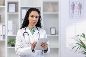 detailopname portret van jong vrouw dokter, Latijns Amerikaans vrouw gebruik makend van tablet computer, op zoek echt nadenkend en gefocust Bij camera, staand binnen kantoor in medisch kantoor van kliniek. foto