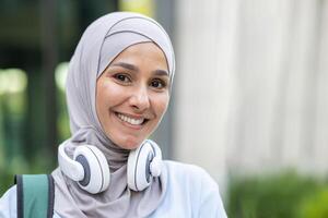 portret van een glimlachen jong moslim vrouw vervelend een hijab en hoofdtelefoons in de omgeving van haar nek, uitdrukken geluk in een stedelijk instelling. foto