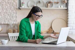 niet succesvol online winkelen. de bank heeft geblokkeerd de account Aan de credit kaart gehouden door een jong vrouw in bril en een groen shirt. ze zit met een laptop Bij huis, boos, verward, verspreidt zich haar handen. foto