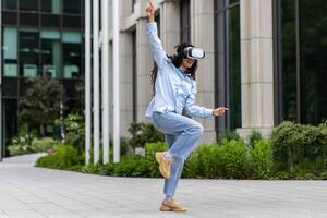 jong mooi meisje in overhemd en jeans dansen buiten kantoor gebouw vervelend virtueel realiteit bril, spaans vrouw gelukkig hebben pret en ontspannende met gekruld haar. foto