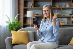 een senior vrouw Aan de sofa proost met een gebalde vuist, op zoek Bij haar smartphone, belichamen vreugde en succes. foto