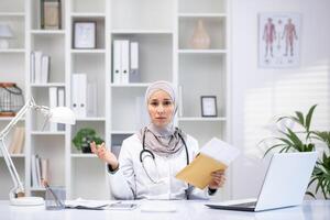 professioneel vrouw in hijab en laboratorium jas in een kliniek kantoor, innemend in geduldig overleg met laptop en medisch apparatuur. foto
