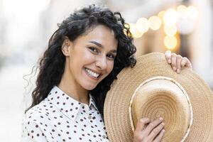 portret van een mooi speels Latijns Amerikaans vrouw, een jong toerist wandelen door de stad in de avond, glimlachen en op zoek Bij de camera, Holding een rietje hoed in haar hand. foto