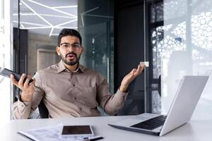 portret van een jong boos en teleurgesteld Indisch Mens zittend Bij een kantoor bureau, Holding een mobiel telefoon en verspreiden zijn handen naar de camera. foto