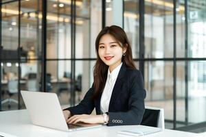 een jong Aziatisch vrouw in een pak zittend Bij een bureau met een laptop in kantoor foto