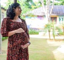 een zwanger Indisch dame poses voor buitenshuis zwangerschap schieten en handen Aan buik, Indisch zwanger vrouw zet haar hand- Aan haar maag met een moederschap jurk Bij maatschappij park, zwanger buiten moederschap schieten foto