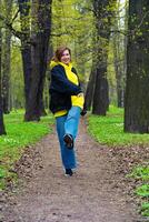 een jong vrouw in een geel capuchon en blauw jeans geniet een ontspannen wandelen in een weelderig groen park gedurende lente, omringd door ontluikend bomen foto