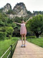 jong vrouw in een roze bloemen jurk genieten van een rustig wandelen Aan een houten brug Bij een berg toevlucht, omringd door weelderig groen foto