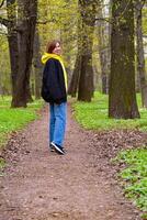 een jong vrouw in een geel capuchon en blauw jeans geniet een ontspannen wandelen in een weelderig groen park gedurende lente, omringd door ontluikend bomen foto