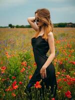 mooi jong meisje in een zwart avond jurk en zonnebril poseren tegen een papaver veld- Aan een bewolkt zomer dag. portret van een vrouw model- buitenshuis. regenachtig het weer. grijs wolken. foto