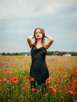 mooi jong meisje in een zwart avond jurk poseren tegen een papaver veld- Aan een bewolkt zomer dag. portret van een vrouw model- buitenshuis. regenachtig het weer. grijs wolken. foto