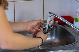 meisje het wassen gerechten in wastafel in camping keuken. jong vrouw is Holding vorken en lepels terwijl spoelen hen. schoonmaak messen en vorken. water is vloeiende en spetterend met spatten uit van tik. foto