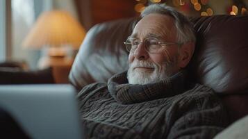 een senior Mens lounges in zijn favoriete fauteuil laptop gestut Aan zijn ronde net zo hij spreekt met zijn dokter door telehealth dankbaar voor de gemak en comfort van virtueel medica foto