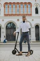 jonge Afro-Amerikaan die mobiele telefoon gebruikt terwijl hij met een elektrische scooter op straat staat foto