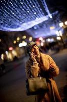 mooie jonge vrouw die haar mobiele telefoon op straat gebruikt in de kersttijd foto