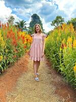 glimlachen jong vrouw in een bloemen zomer jurk staand in een levendig rood bloem veld- met een toneel- berg achtergrond, vastleggen de essence van zomer foto