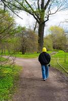 een jong vrouw in een geel capuchon en blauw jeans geniet een ontspannen wandelen in een weelderig groen park gedurende lente, omringd door ontluikend bomen foto