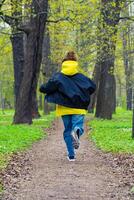 een jong vrouw in een geel capuchon en blauw jeans geniet een ontspannen wandelen in een weelderig groen park gedurende lente, omringd door ontluikend bomen foto