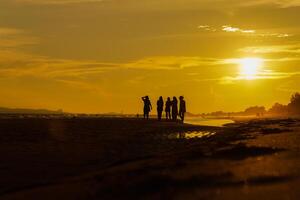 silhouet van een groep van vrienden genieten van de zonsondergang Aan de strand Bij gouden zonsondergang tijd, ontspannende Aan een ontspannende vakantie, zee in de avond. foto