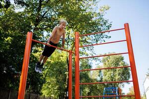 een tiener Aan de dwarsbalk presteert acrobatisch elementen. straat training Aan een horizontaal bar in de school- park. foto