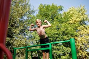 een sport- kind beklommen op de sport- horizontaal bars en shows de spieren van de armen. straat training Aan een horizontaal bar in de school- park. foto