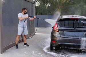 de Mens heeft een water kanon in zijn hand- dat ze toepassingen naar wassen de auto.a auto Bij een zelf onderhoud auto wassen. foto