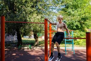 atletisch tiener zit Aan de dwarsbalk, Holding Aan naar de leuning. straat training Aan een horizontaal bar in de school- park. foto