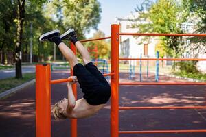 een tiener presteert opdrachten door het werpen zijn poten over- de horizontaal bar. straat training Aan een horizontaal bar in de school- park. foto