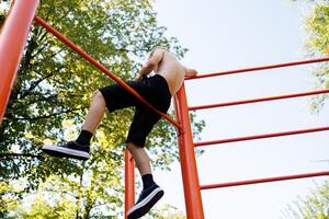 visie van de bodem van een tiener wie zit Aan een gymnastiek- dwarsbalk en horloges. straat training Aan een horizontaal bar in de school- park. foto