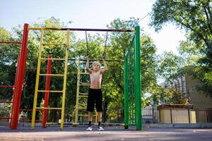 een atletisch kind gepakt de ringen naar uitvoeren een optrekken. straat training Aan een horizontaal bar in de school- park. foto