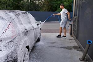 de kant van de auto is gedekt met schuim, de persoon in de achtergrond is toepassen schuim naar de auto. een auto Bij een zelf onderhoud auto wassen. foto